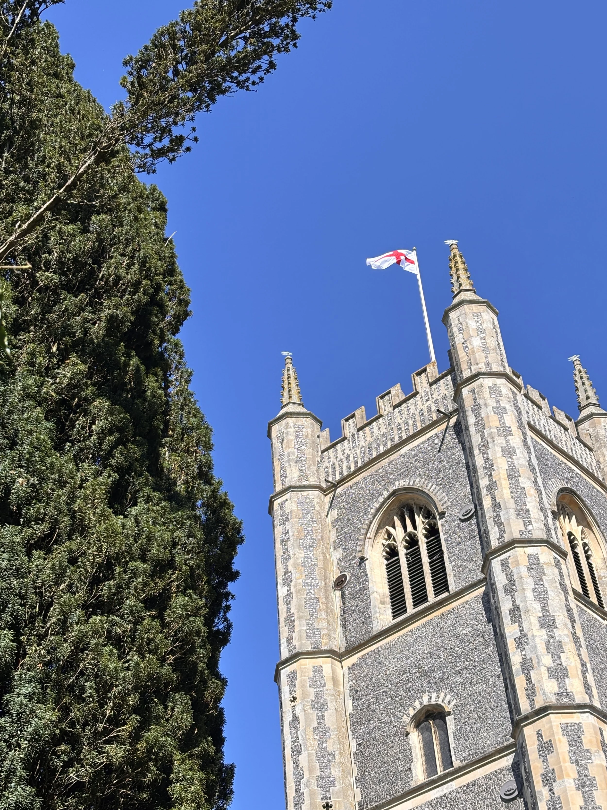A tall church tower made of stone and brick with pointed turrets, flying the England flag, and surrounded by green trees under a clear blue sky.