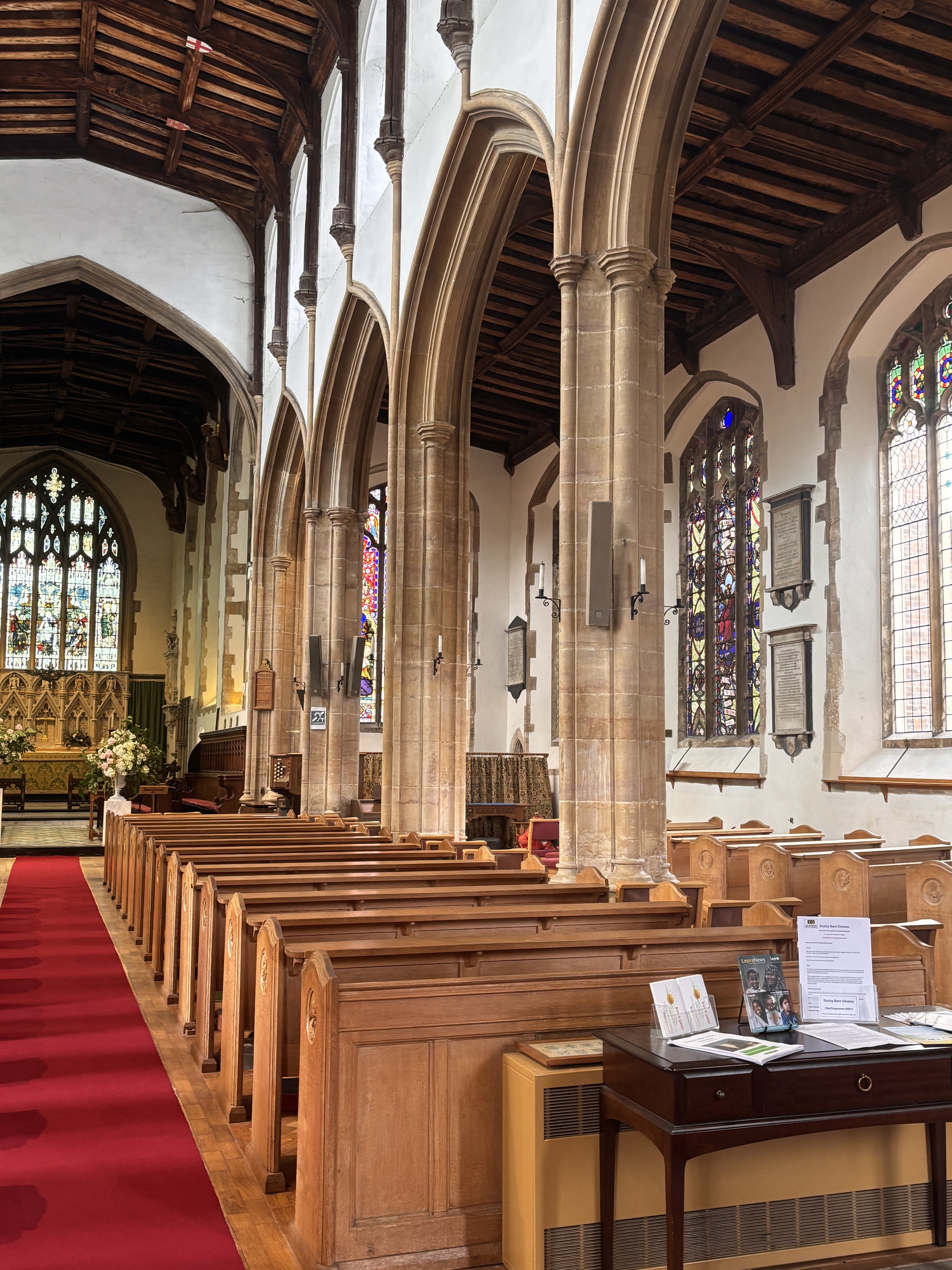 Interior of a church with wooden pews, stone columns, stained glass windows, and an altar area with flowers and religious decorations.