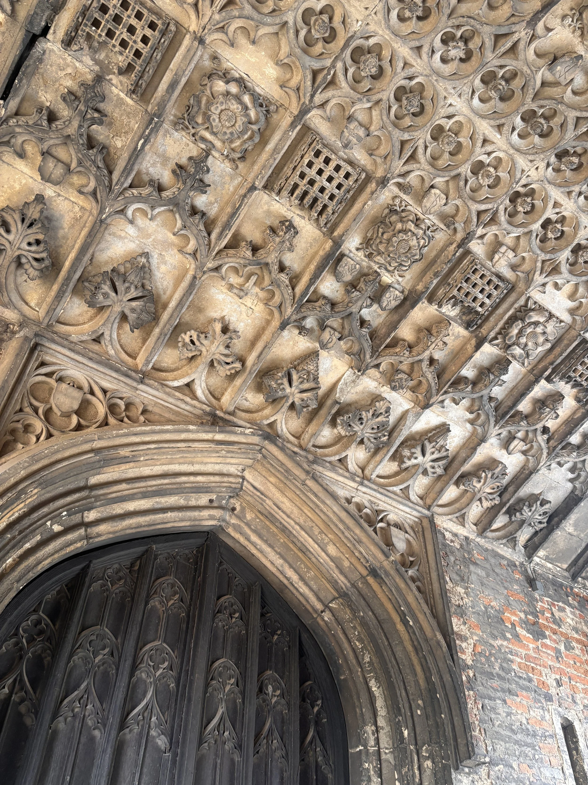 Ornate Gothic-style archway with detailed stone carvings, floral patterns, and decorative vents, leading to wooden doors in an old building.