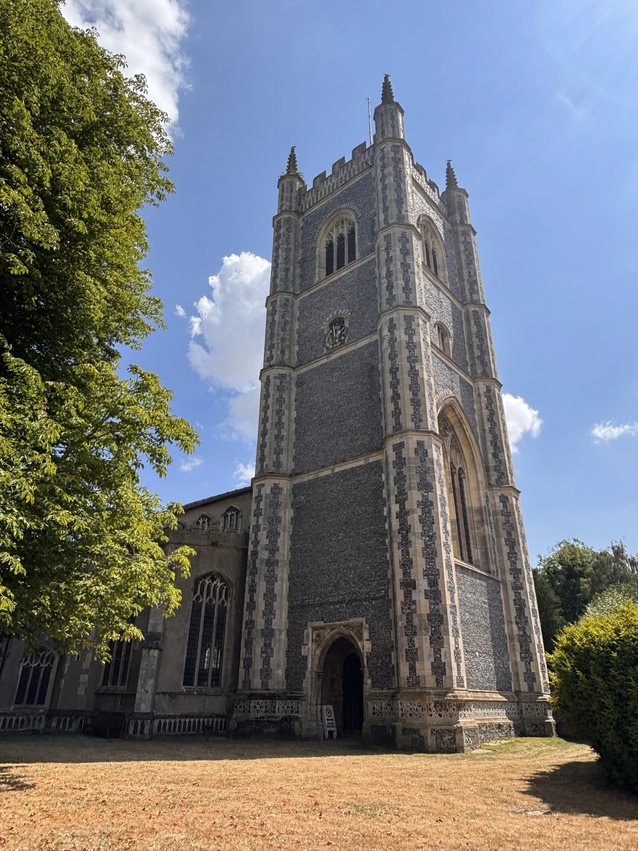 Exterior of an old stone church with tall clock tower, surrounded by trees and a grassy area in the foreground.