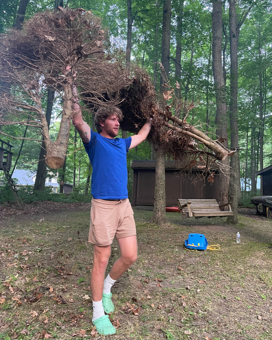 A young man wearing a blue t-shirt, beige shorts, white socks, and green Crocs is lifting a large uprooted tree branch in a wooded backyard. There is a blue toolbox and a water bottle on the ground nearby, with trees and a shed in the background.