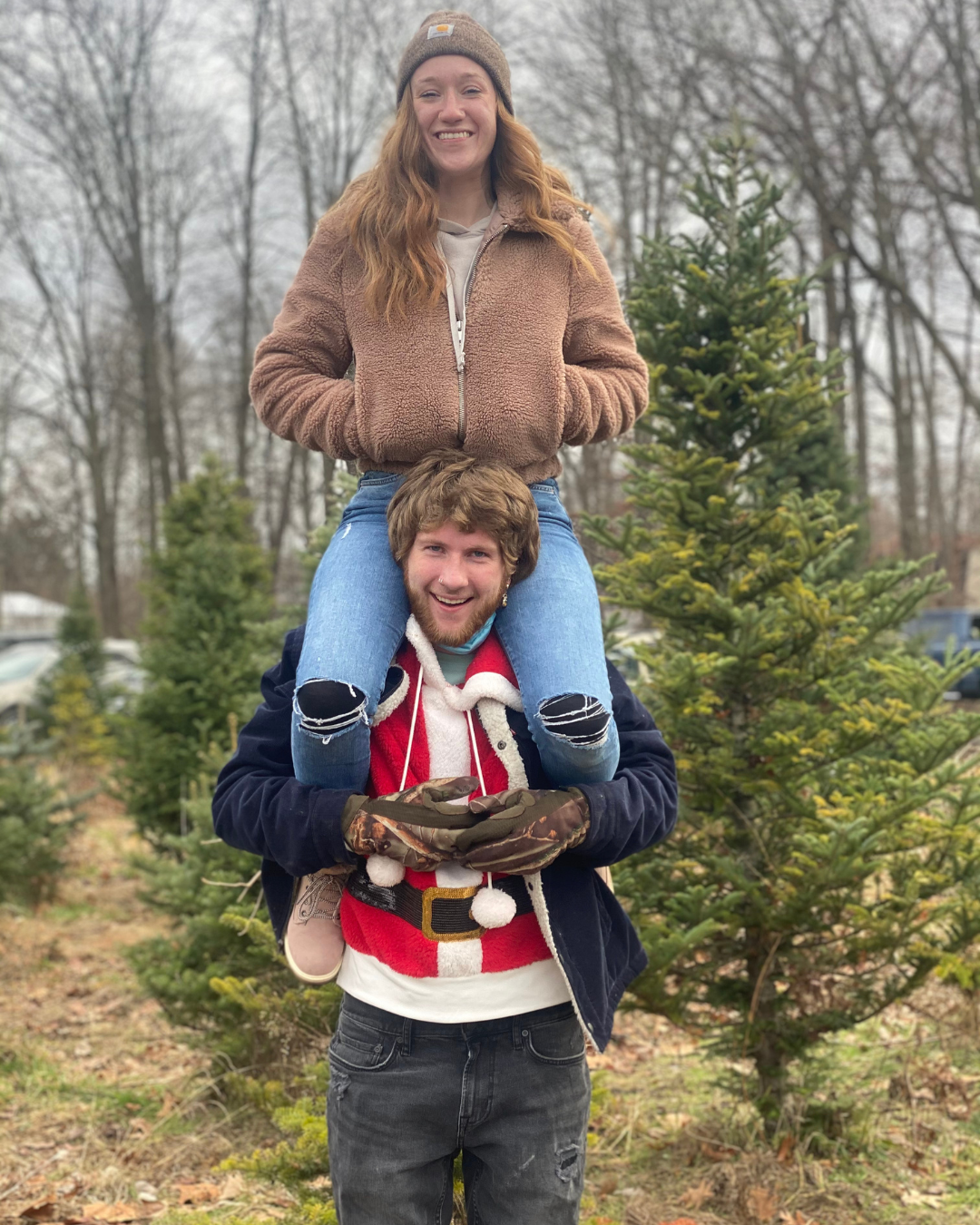 A woman wearing a beanie hat and a brown fleece jacket sitting on a man's shoulders, with her hands in her jacket pockets, smiling at the camera. The man is wearing a Santa Claus-themed sweater and camouflage gloves, carrying her on his shoulders. They are outdoors in a Christmas tree farm or forest area, with several small Christmas trees and leafless trees in the background.