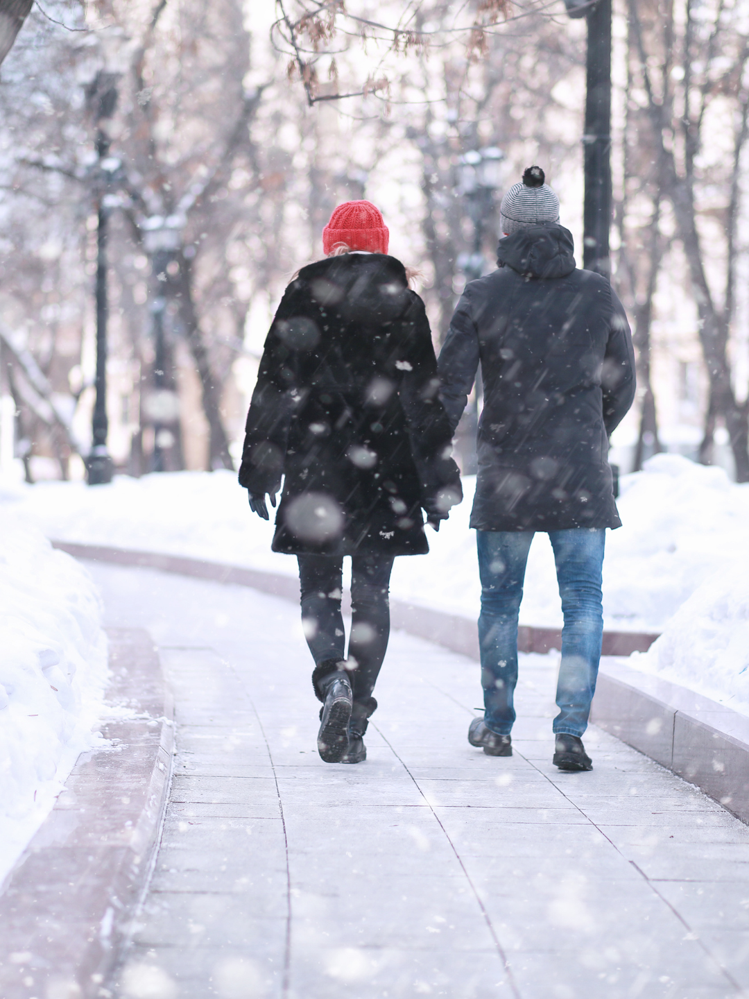 Two people wearing winter coats and hats walking hand-in-hand on a snowy path in a park during snowfall.