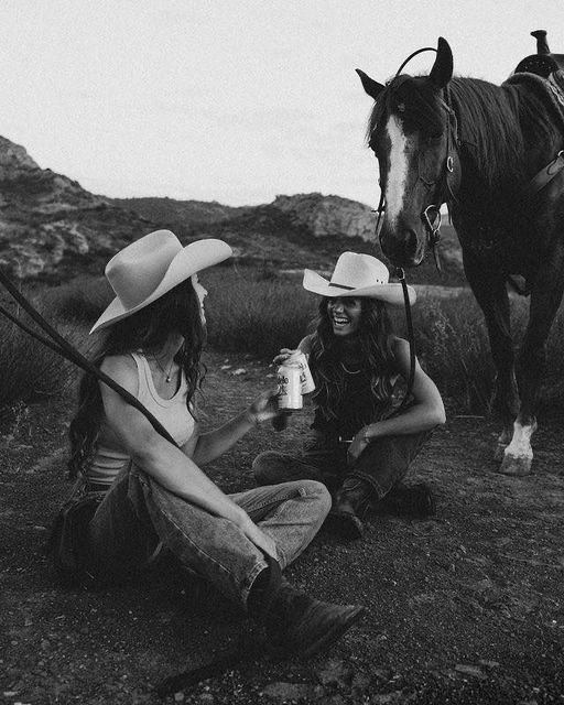 Two women wearing large cowboy hats sitting on the ground and smiling, with a horse standing nearby in a rural outdoor setting.