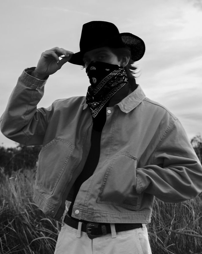 A person wearing a cowboy hat, bandana over the face, denim jacket, and light pants standing outdoors in a field with tall grass, with an overcast sky.