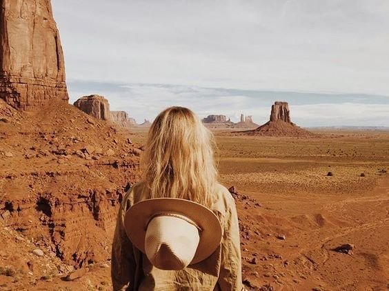 A woman with blonde hair standing in a desert landscape with large rock formations and mesas in the distance, wearing a hat around her neck.
