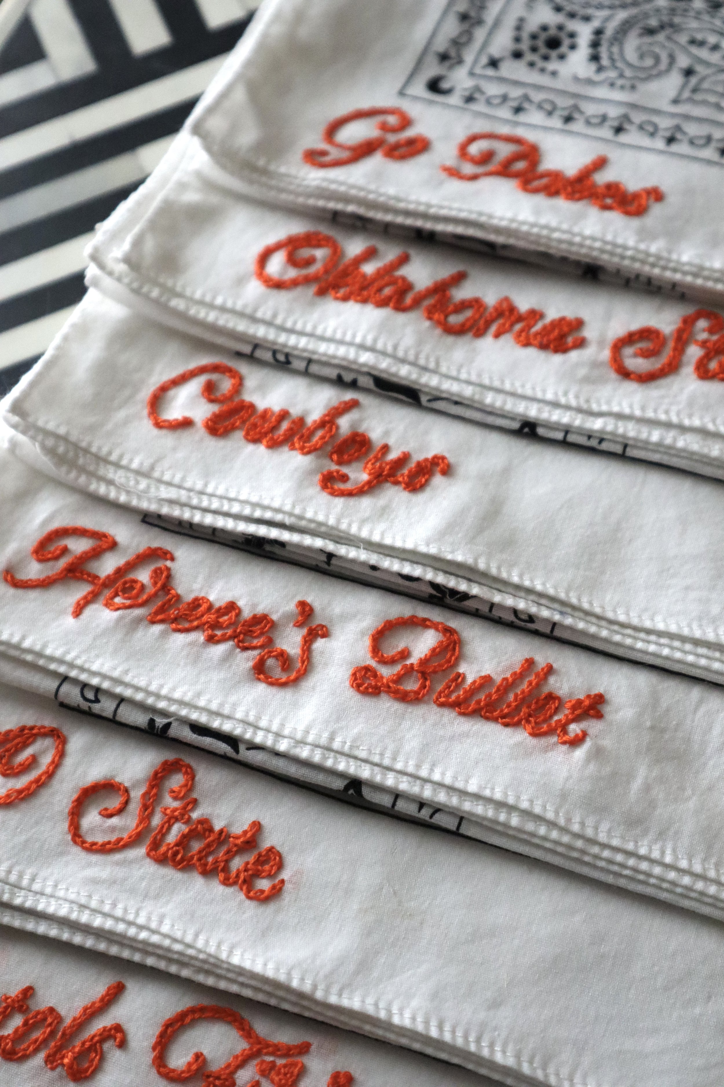 Close-up of white cloths with red embroidered text that says 'Go Dad's or' and other words partially visible. Each cloth appears to be folded and layered on a black and white striped surface.