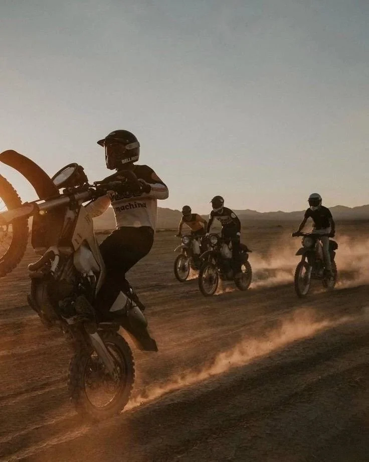 Four dirt bike riders riding on a dusty trail in a desert landscape during sunset, with mountains in the background.