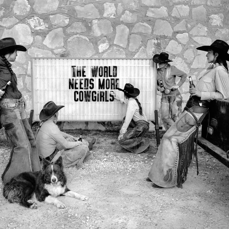 Group of women dressed as cowgirls with hats, jeans, and boots, gathered around a sign that reads "The world needs more cowgirls". One woman is sitting on the ground, another is kneeling, and the rest are standing, with a dog lying on the ground nearby, and a saddle on a rack.
