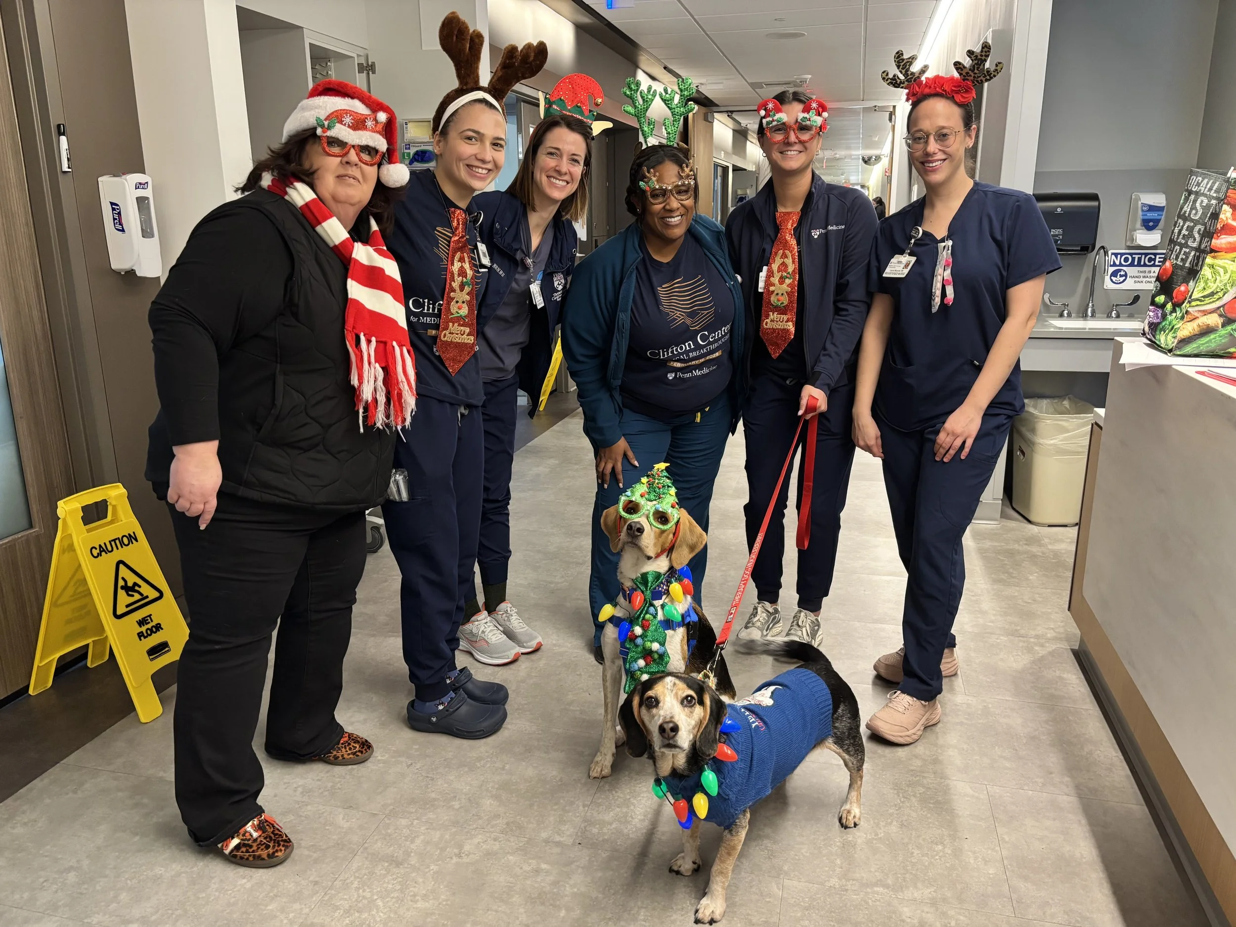 Six women and three dogs dressed in festive holiday costumes and accessories standing in a hospital corridor.