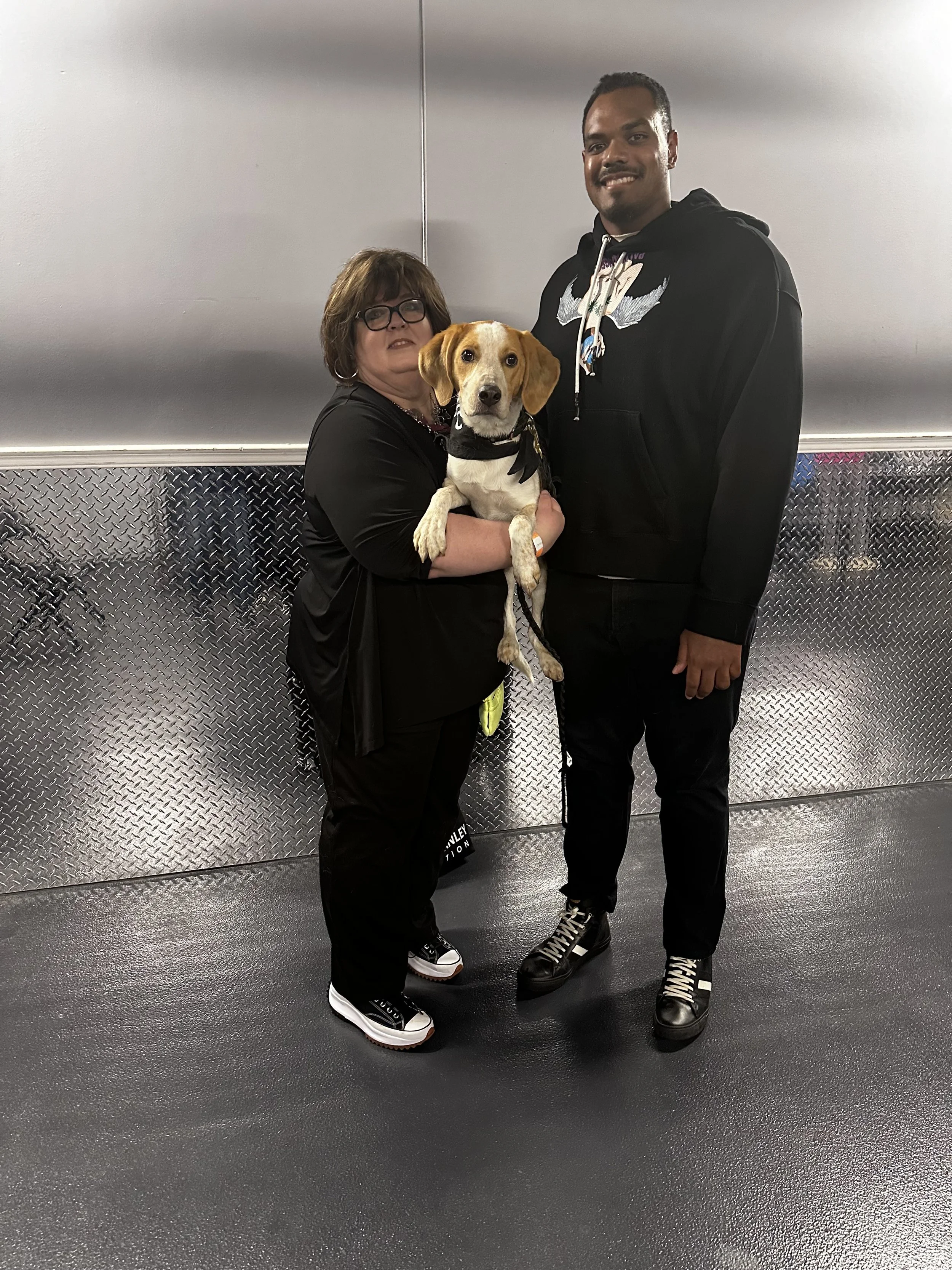 A woman with glasses holding a tan and white dog, standing next to a tall man in a black hoodie, inside a workshop or garage with a metallic wall background.