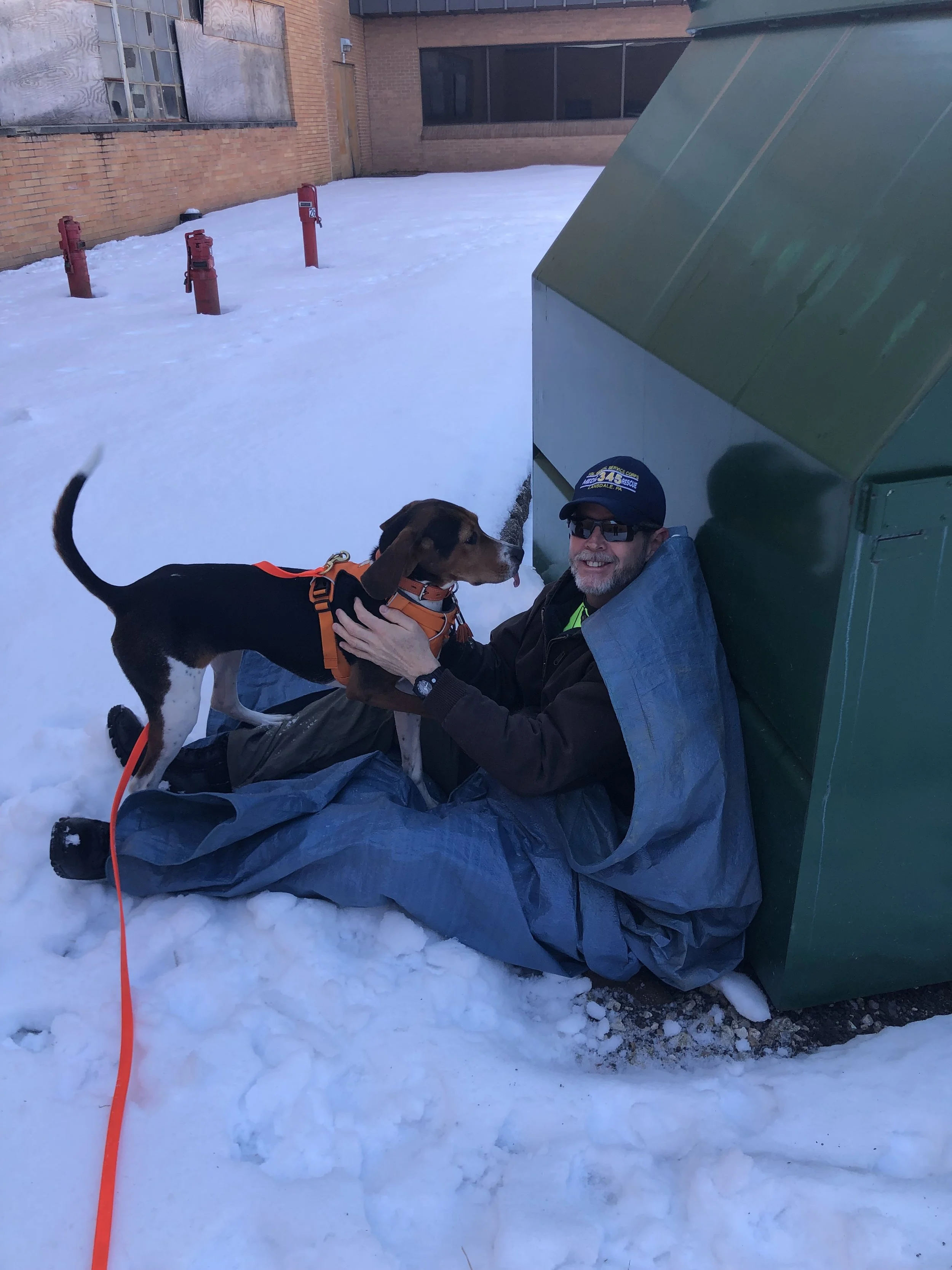 A man and a dog are sitting on snow-covered ground outside a brick building. The man is smiling, wearing sunglasses, a cap, and a gray jacket. The dog is a hound, wearing an orange harness, and is interacting with the man. There is a green utility box or shed to the right, and three red fire hydrants are visible in the background.