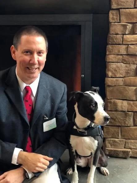 A man in a suit sitting next to a black and white dog in front of a fireplace and brick wall.