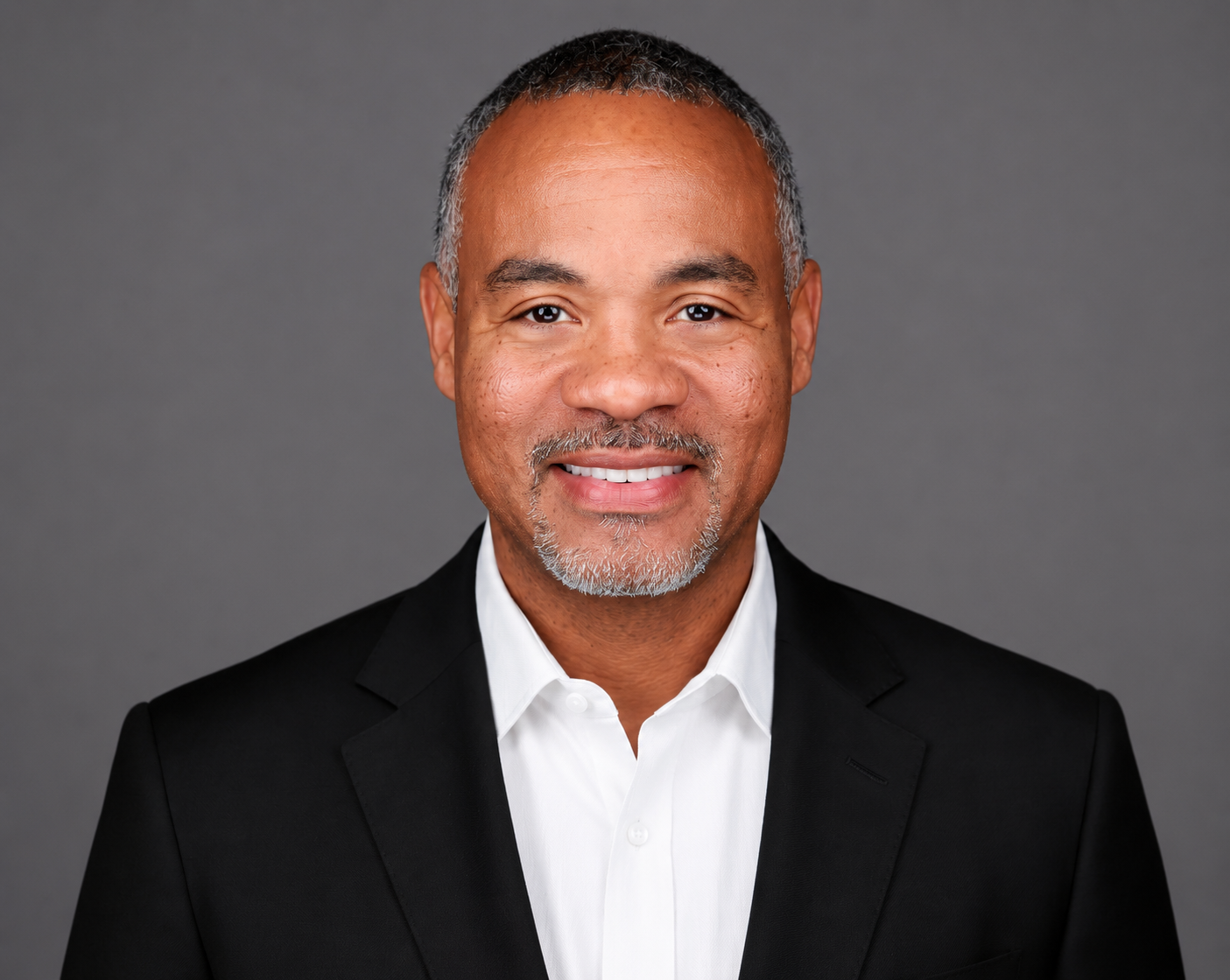 A professional headshot of a middle-aged man with short gray hair, a gray beard, wearing a black suit jacket and white dress shirt, smiling against a gray background.