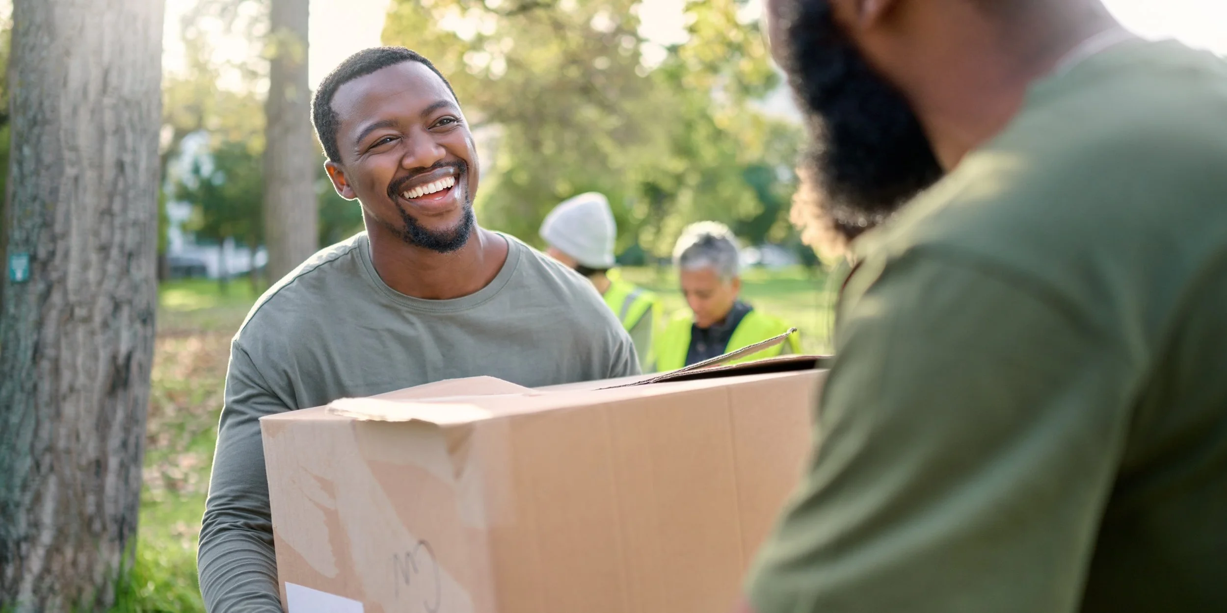A smiling man in a gray shirt hands over a cardboard box to a person with a beard wearing a green shirt. They are in a park with trees and sunlight. Two people in yellow vests are in the background.