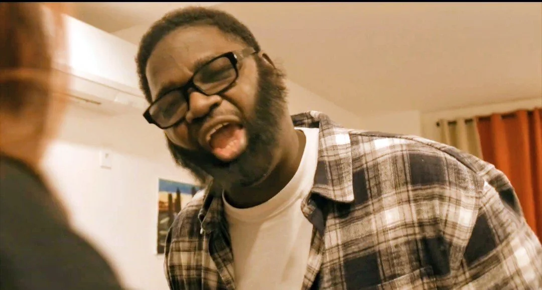 A man with glasses, facial hair, and wearing a plaid shirt, appears to be yelling or talking loudly, inside a home with curtains and a picture or poster on the wall.