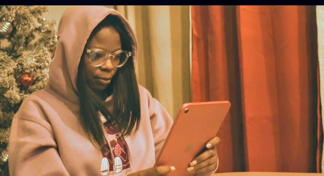 Erica is wearing a lavender hoodie and glasses while looking at her tablet at the dinner table.