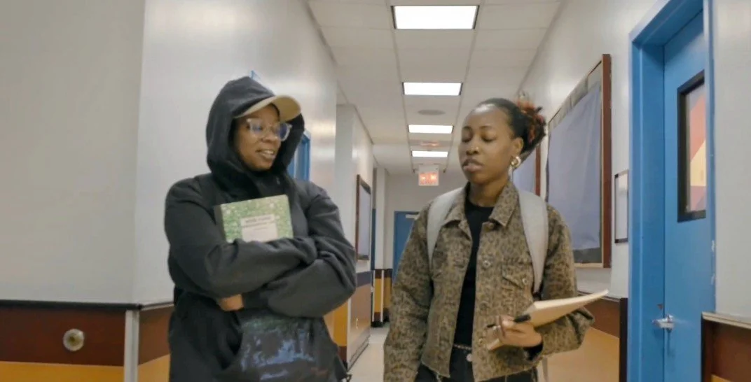 Two students walking down a school hallway, one holding a notebook, the other a folder, engaged in conversation.