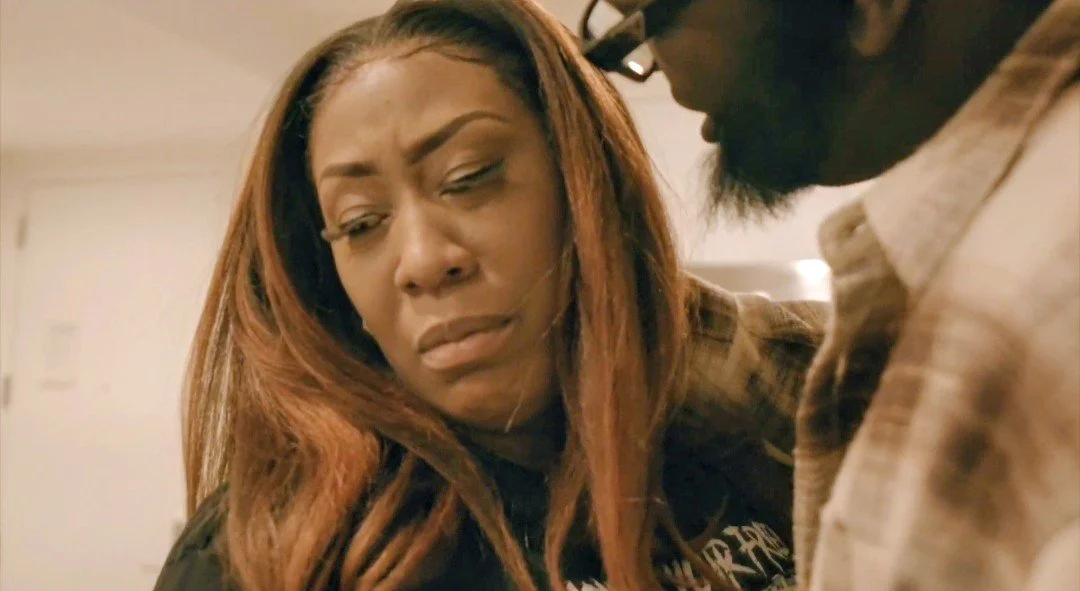 A woman with long reddish-brown hair looks upset and is close to a man who has dark skin, glasses, a beard, and is wearing a plaid shirt. They appear to be in an indoor setting.