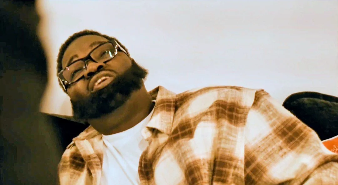 A man with glasses and a beard wearing a plaid shirt, sitting and looking toward the left.
