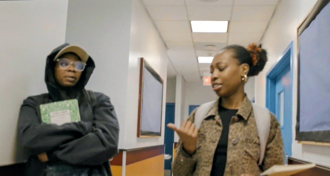 Two women talking in a school hallway, one with a backpack and the other holding a notebook.
