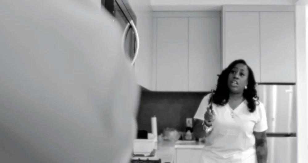 A woman with dark, curly hair standing in a modern kitchen holding a utensil, with kitchen cabinets and appliances visible in the background.