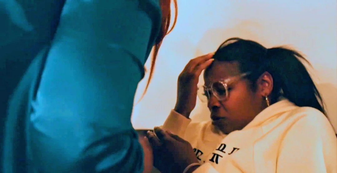A woman with long dark hair, glasses, and hoop earrings appears distressed as another person, whose face is not visible, touches her arm. She is wearing a white hoodie and is sitting against a plain wall.