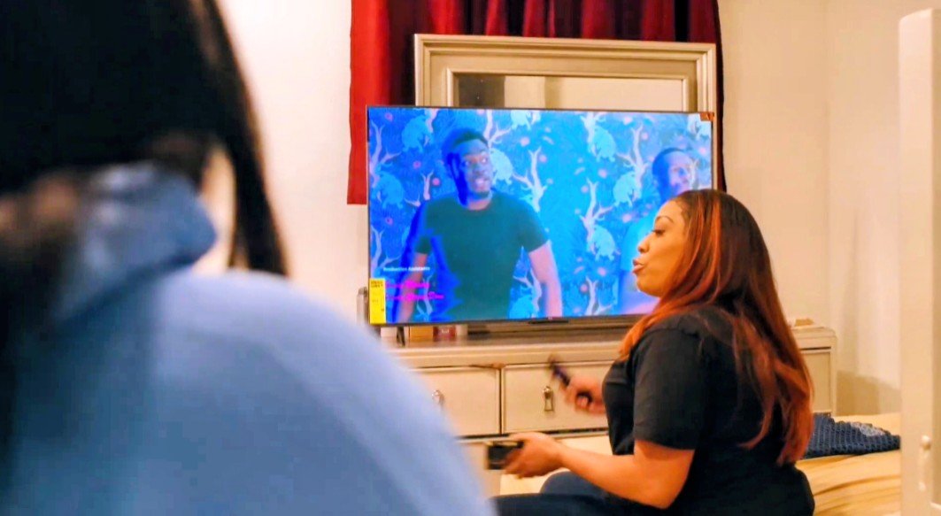 Two women watching a television screen with a man on it in a living room, with a mirror above the TV and red curtains.