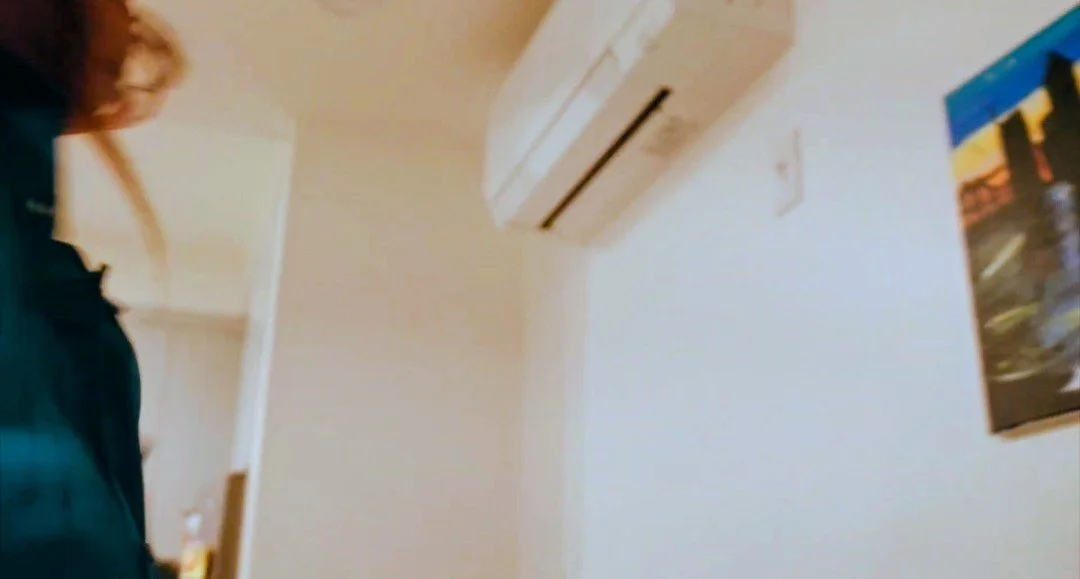 A person with red hair and wearing a dark-colored shirt standing in a room with white walls and an air conditioning unit on the ceiling. A poster is partially visible on the wall.