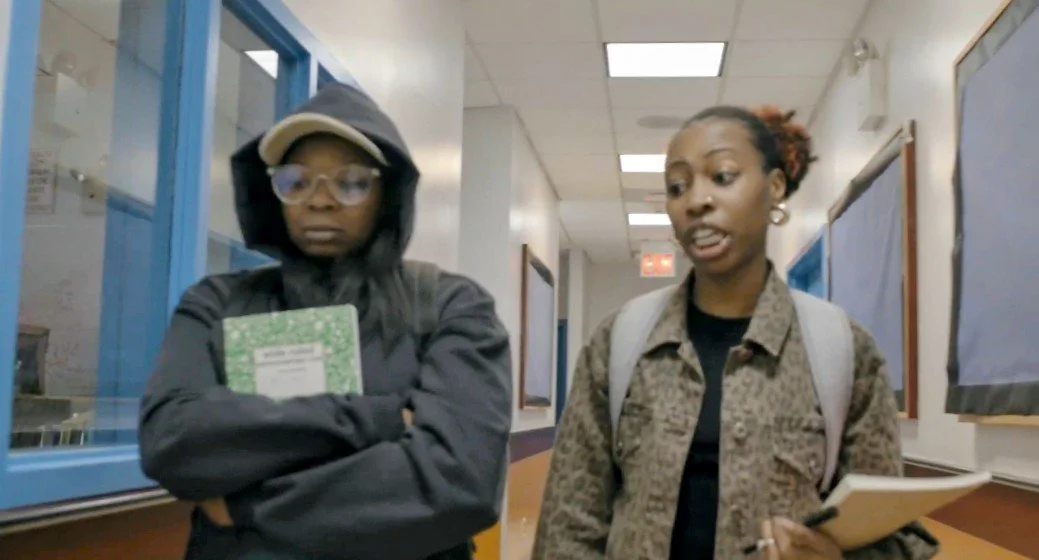 Two teenage students talking in a school corridor. One student is wearing glasses, a hoodie, and holding a notebook, with arms crossed. The other student is speaking, wearing a patterned jacket and carrying a backpack and notebook.