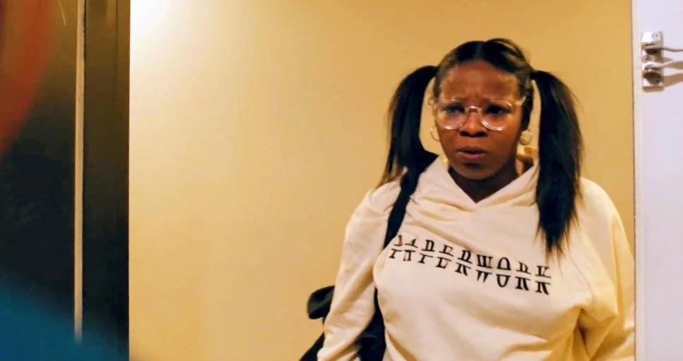 A woman with pigtails, wearing glasses, headphones, and a cream hoodie with text, appears surprised or confused while standing in a doorway.