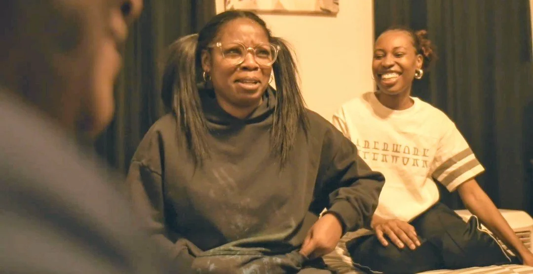 Two women sitting and laughing in a room, with a dark curtain behind them. One woman is wearing glasses and a dark hoodie, the other is wearing a white t-shirt with text on it.