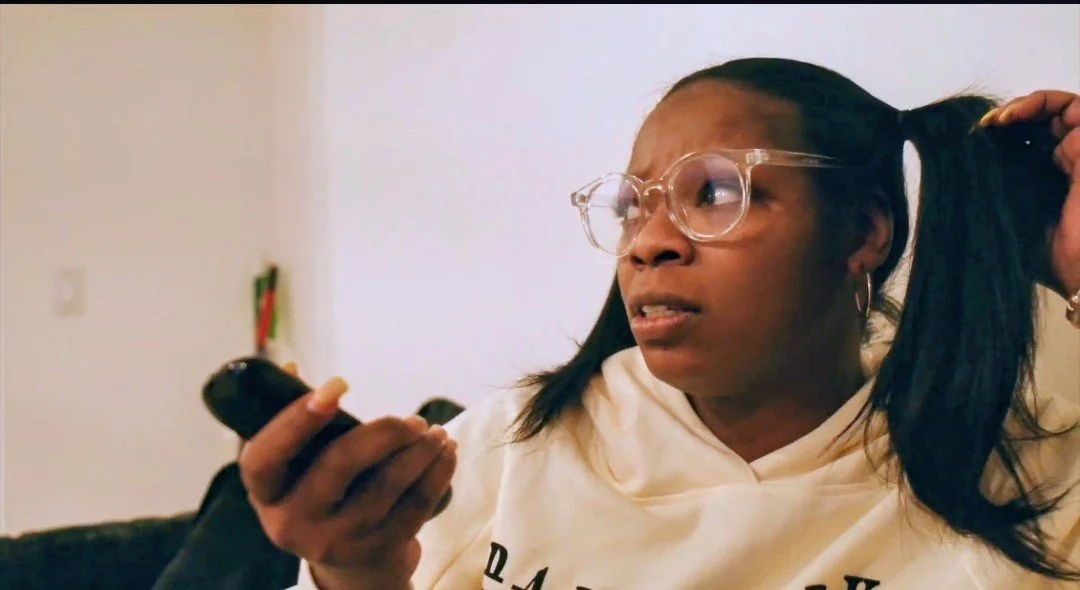 A woman with glasses and hoop earrings holding a phone in her right hand, untangling or adjusting her long hair with her left hand, indoors with a plain wall behind her.