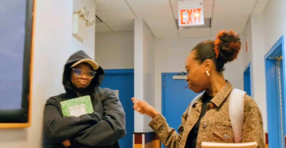 Two women having a conversation in a hallway near blue doors. One woman is leaning against the wall with arms crossed, wearing a hoodie and glasses. The other woman is gesturing with her finger, wearing a patterned jacket and a backpack.