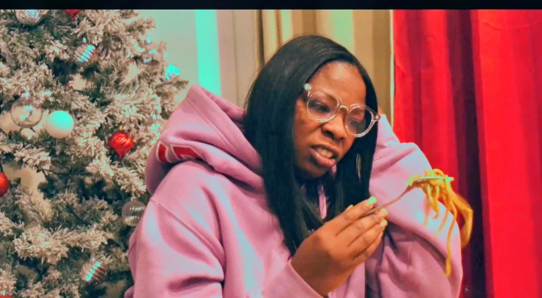 A woman with long black hair, glasses, wearing a pink hoodie, eating spaghetti with a fork, standing near a decorated Christmas tree with red and white ornaments.