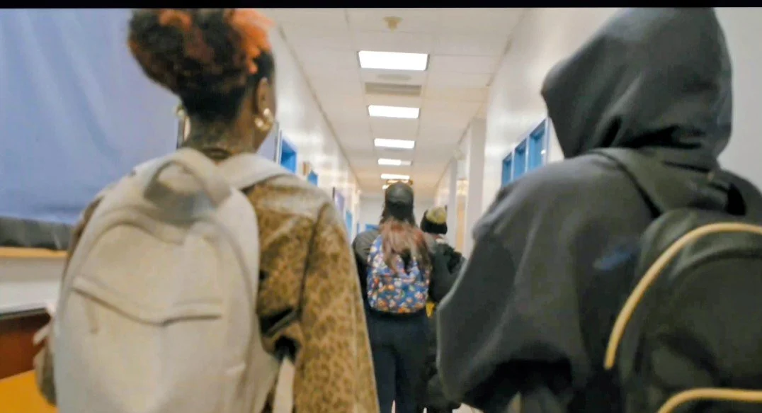 Students walking in a school hallway, carrying backpacks, some wear hoodies, others wear jackets, with lockers on both sides of the hallway.