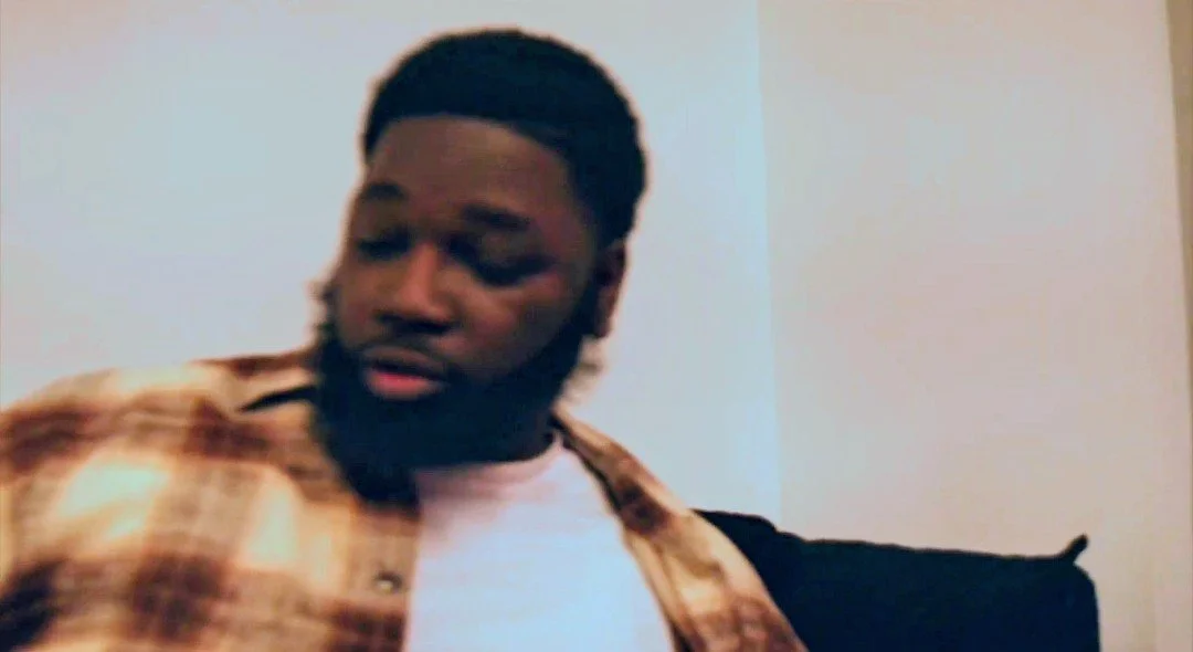 A man with dark skin, a beard, and short hair styled in a fade, wearing a plaid shirt over a white t-shirt, looking down or to the side inside a room with plain walls.
