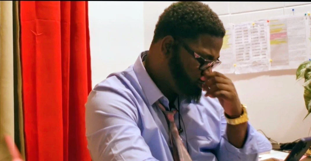 A man with glasses, a beard, and short curly hair, wearing a light blue shirt and a tie, sitting at a desk, holding his nose with his right hand, with a wall with papers and a plant in the background.