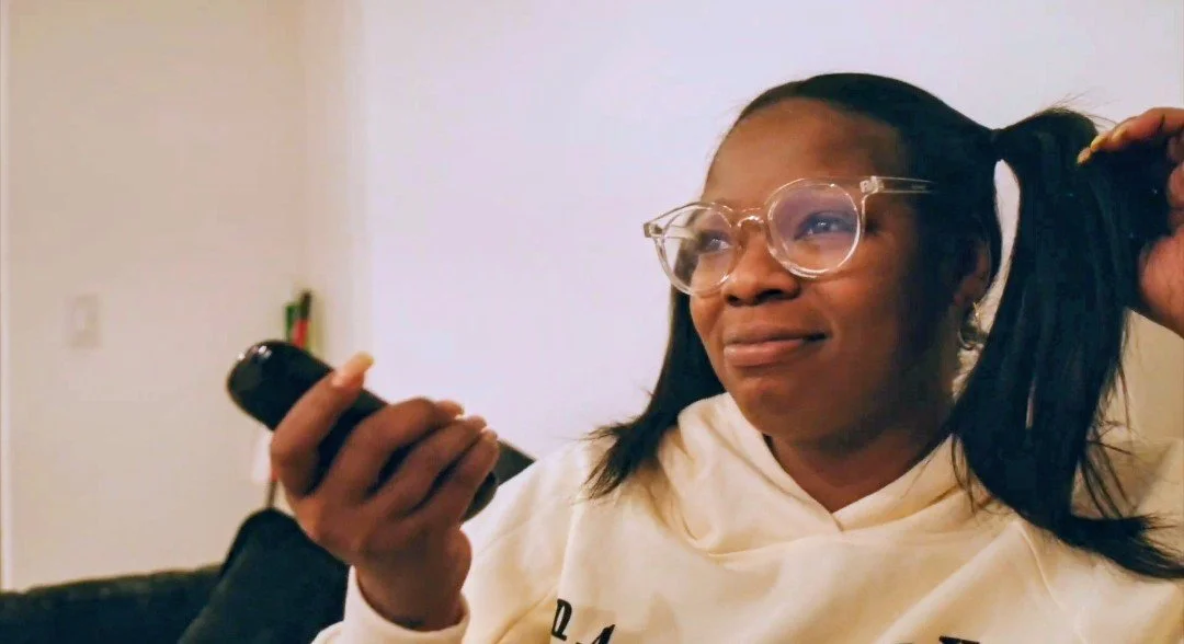 A woman with glasses and pigtails looking at her phone, sitting on a couch in a home interior.