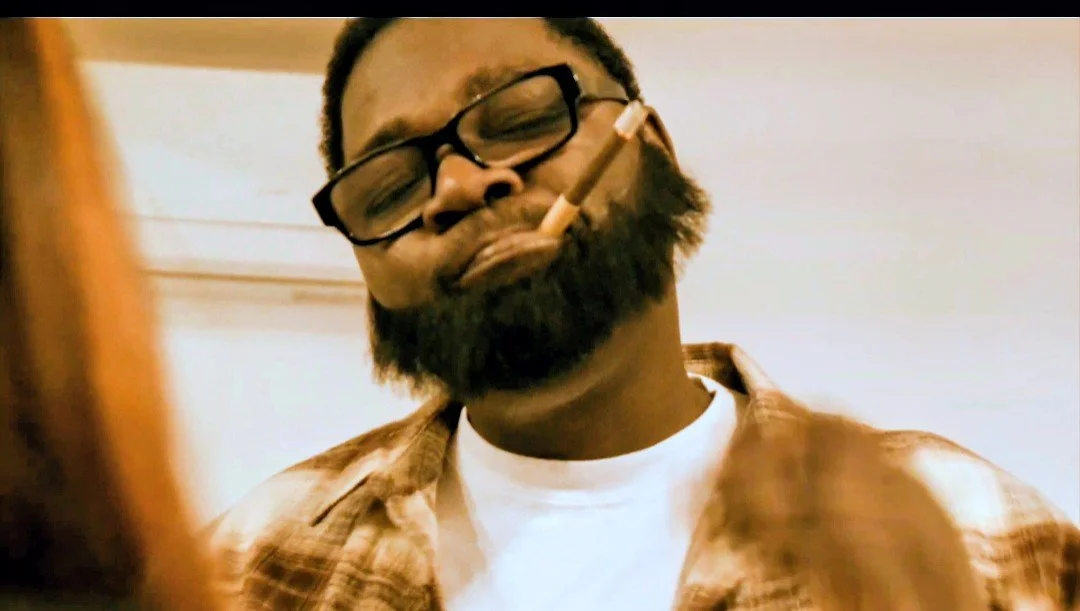 A man with a beard and glasses holding a cigarette in his mouth and wearing a plaid shirt and a white T-shirt, with a blurred foreground.