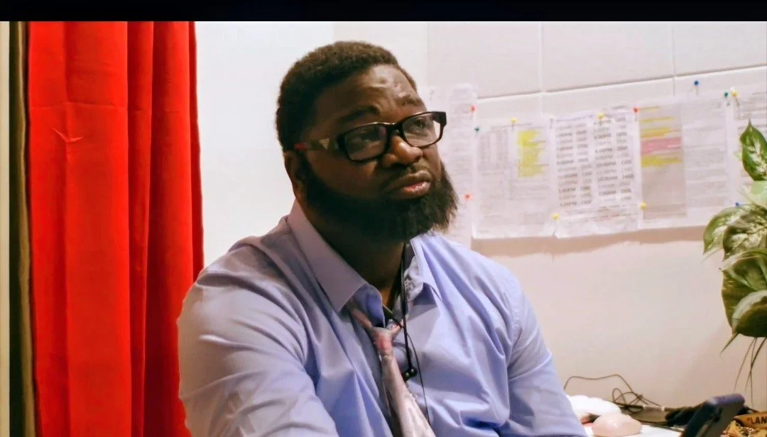 A man with glasses and a beard, wearing a light blue shirt and a pink tie, sitting in an office with white walls, red curtains, and papers pinned to the wall behind him.