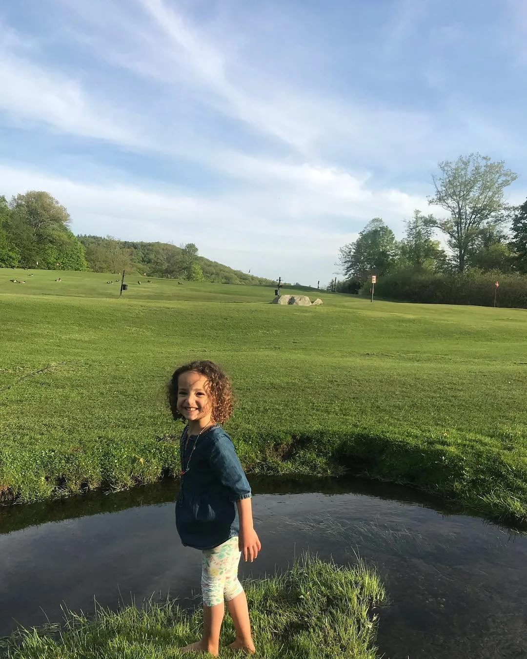 A young girl standing in a small pond or water feature on a grassy field with a golf course and trees in the background under a blue sky with some clouds.