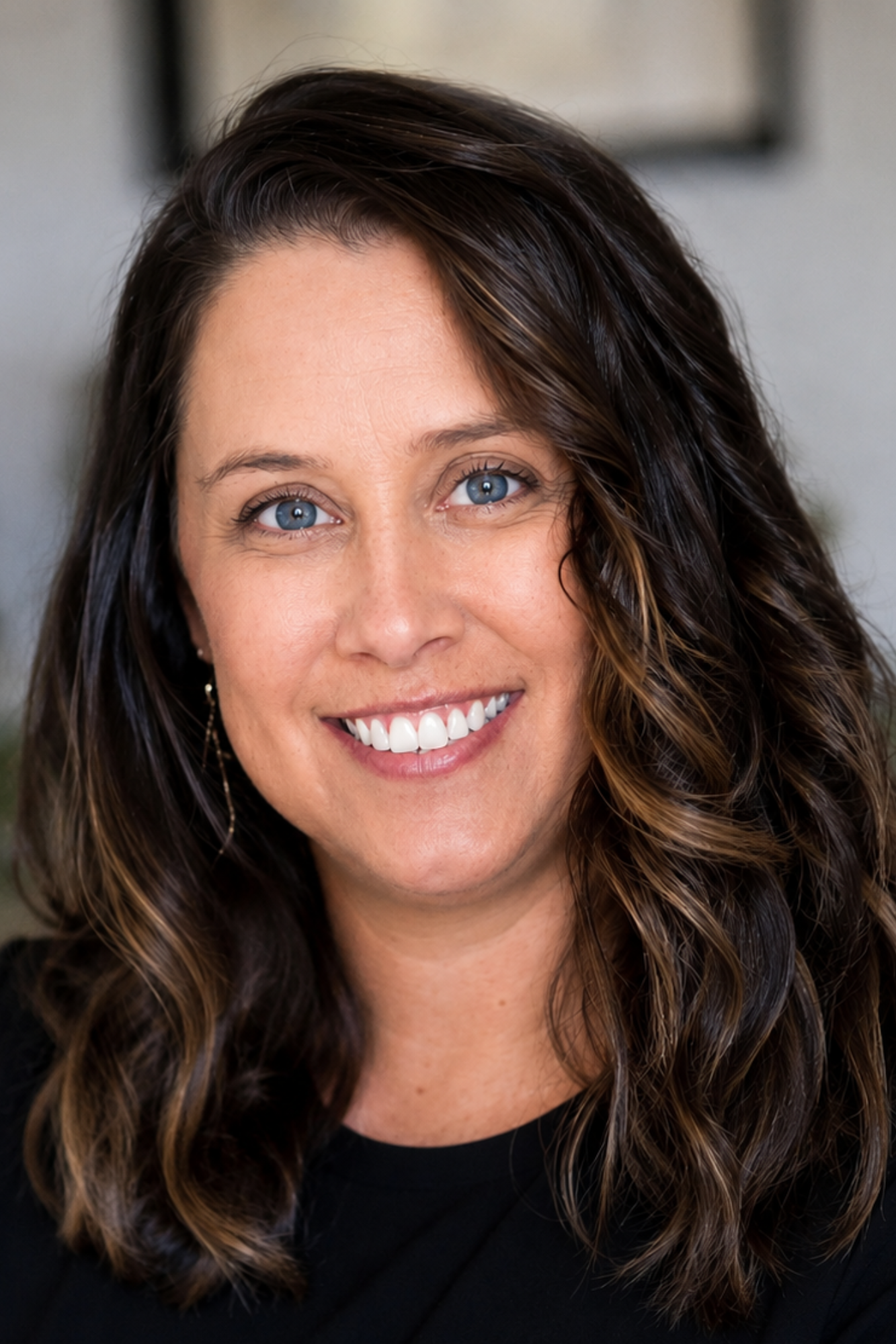 A close-up of a woman with curly, dark hair with lighter streaks, blue eyes, and a broad smile showing white teeth, wearing earrings and a black top, bright indoor lighting.