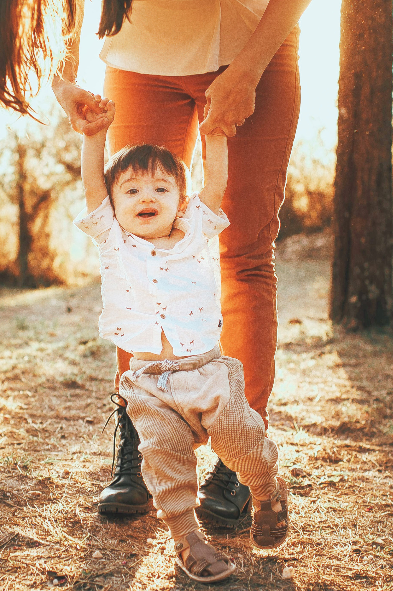 A young child with dark hair and light skin is holding hands with an adult while walking outdoors on a sunny day. The child is smiling and wearing a white button-up shirt, tan pants, and sneakers. The adult is wearing orange pants and a light-colored shirt. There are trees and sunlight in the background.