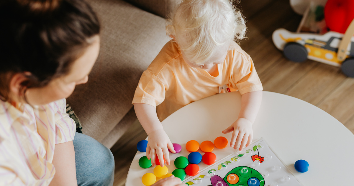 Two children playing a colorful bubble popping game on a white table, with a brown sofa and a wooden floor in the background.