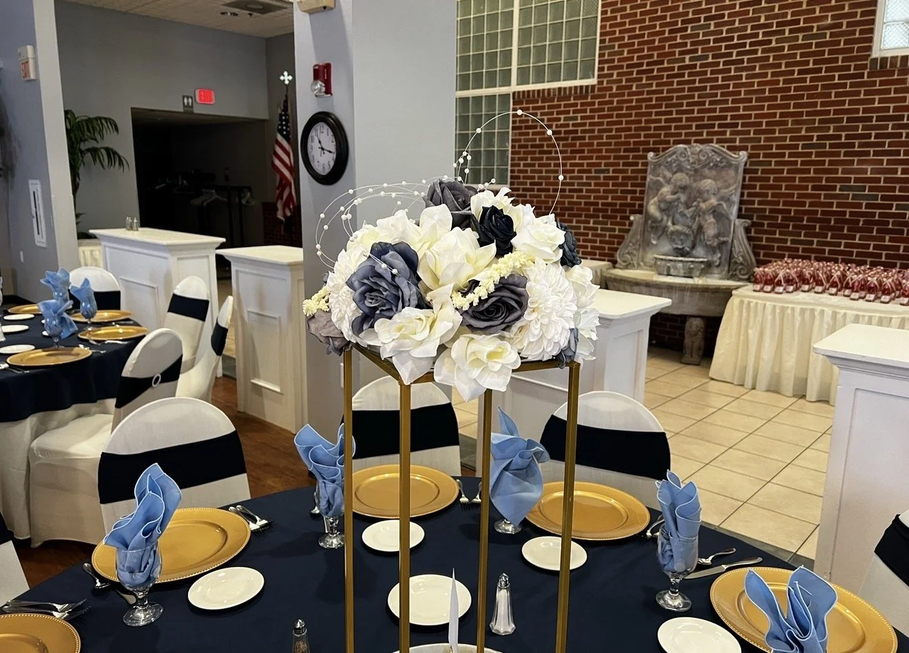 A wedding reception table with black tablecloth, gold plates, and blue cloth napkins. A floral centerpiece with white, gray, and black flowers is displayed on a taller stand. In the background, there are other tables, a wall with brick and glass bloc