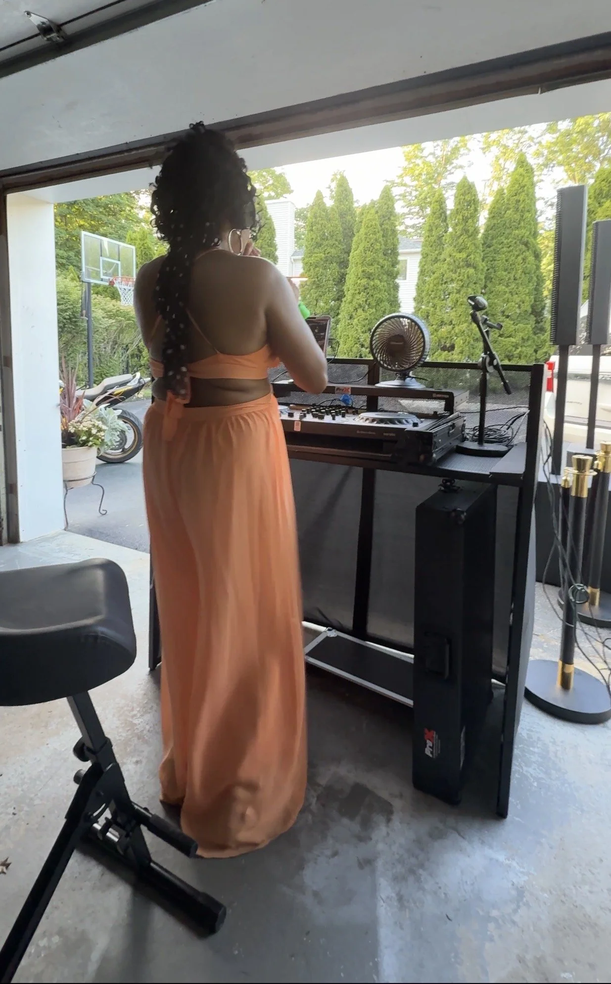 A woman with curly hair wearing a peach-colored, strapless top and matching long skirt, standing in a garage or open-air space, using a DJ mixer or controller on a table. There is a fan, a microphone stand, and two large speakers nearby, with greener