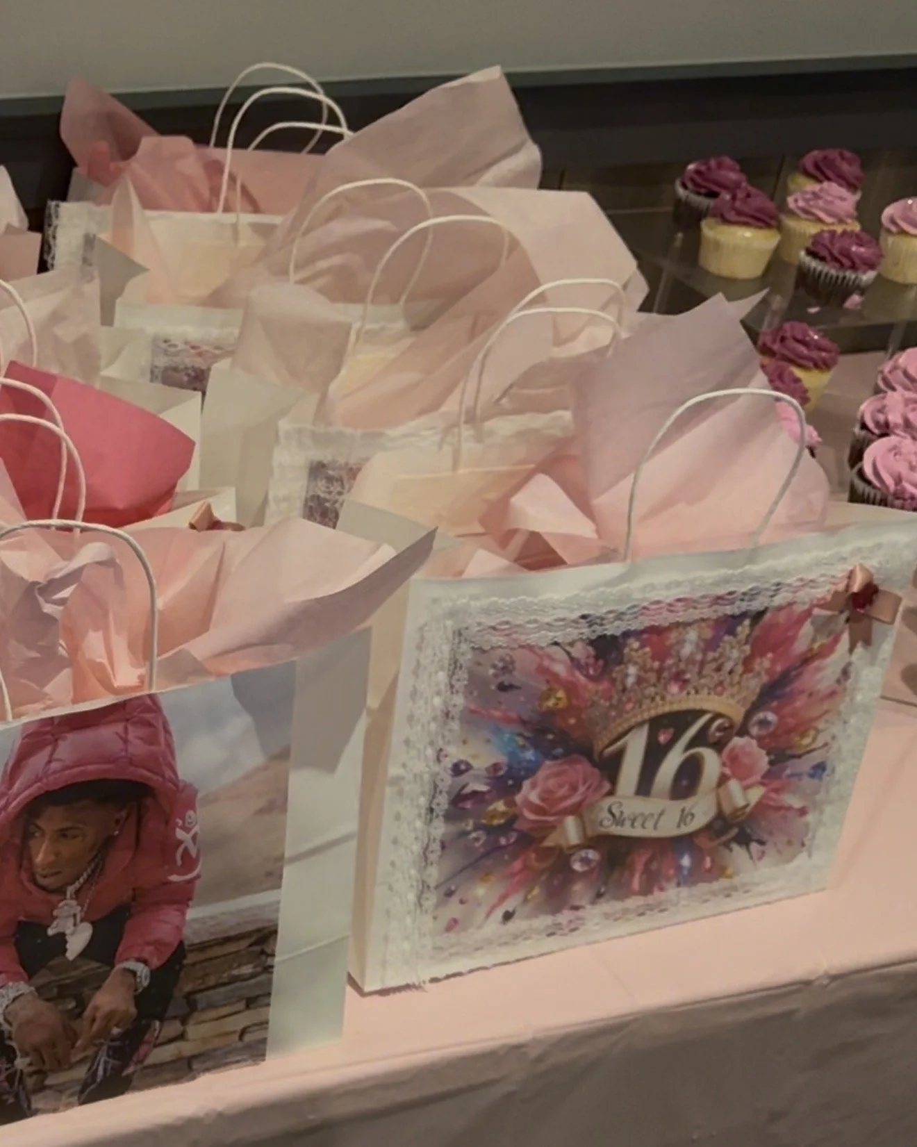 Pink gift bags with tissue paper and a pack of cupcakes with pink frosting on a table.