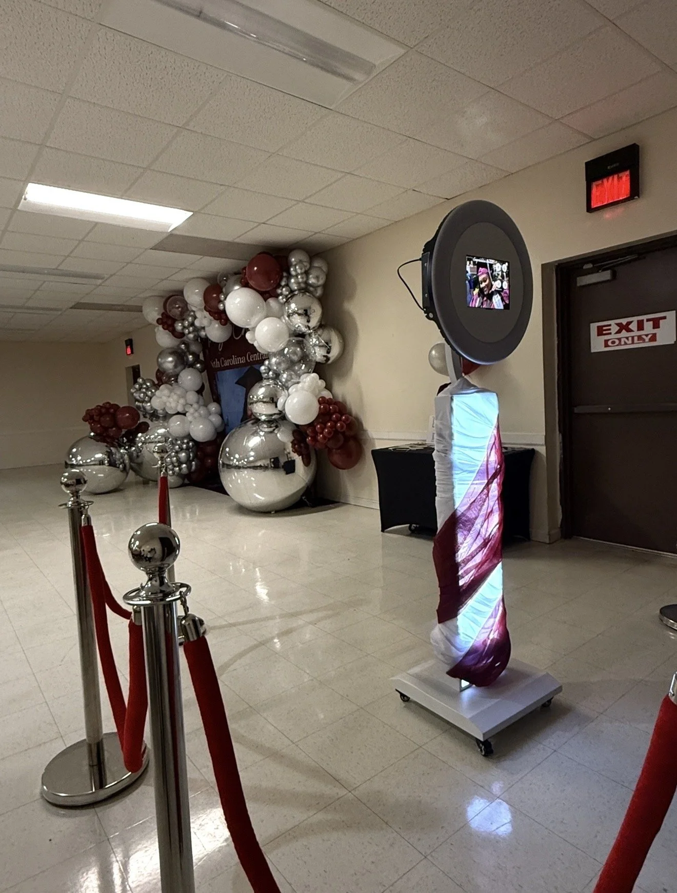 Decorative balloon arrangement with red, white, and silver balloons and an American flag-themed drape at an indoor event space, with a red velvet rope barrier in the foreground and a large speaker display to the right.