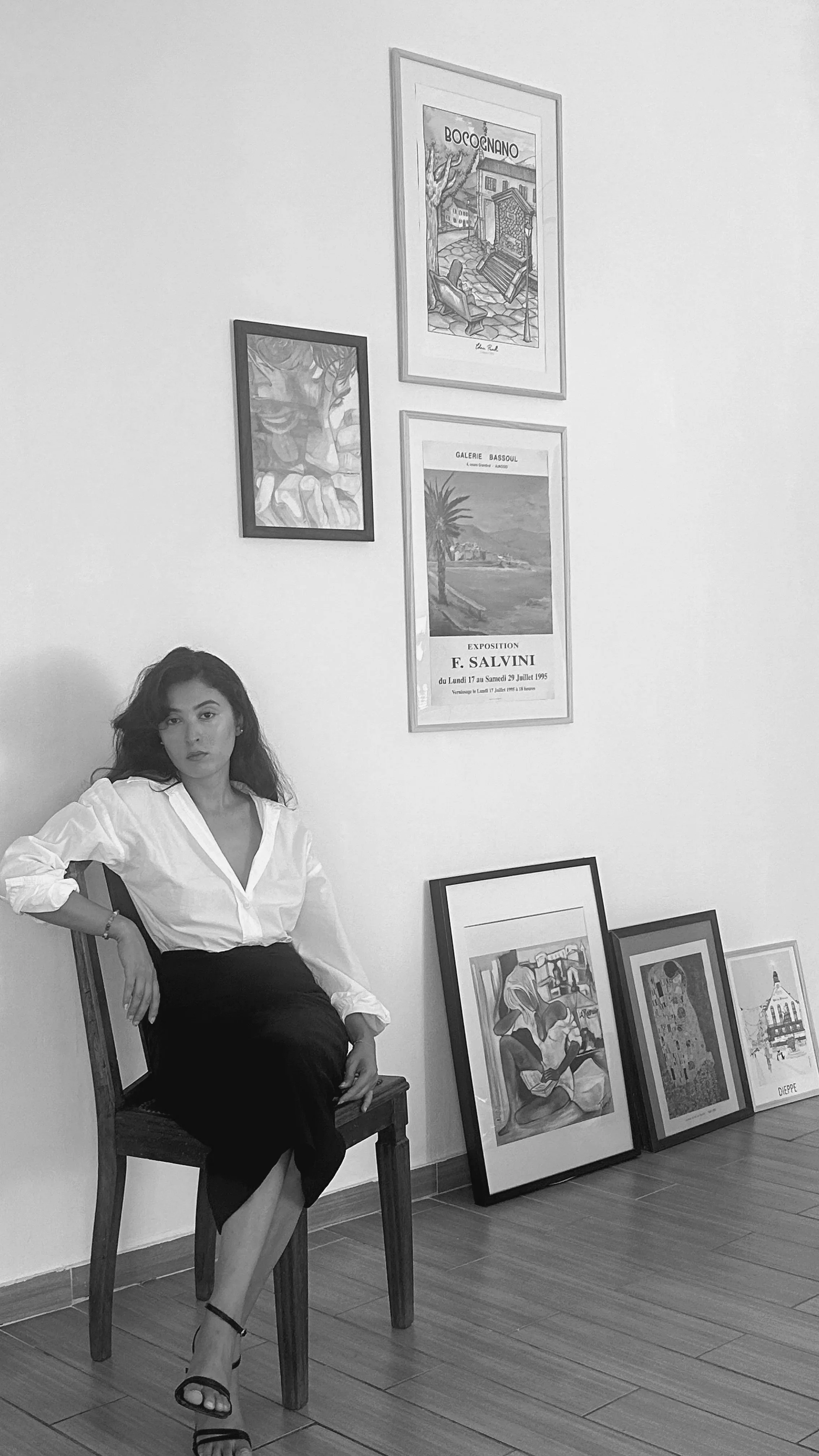 Une femme élégante assise sur une chaise en bois dans une galerie d'art, entourée de plusieurs œuvres d'art suspendues au mur et posées par terre.